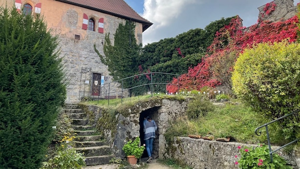 A stone building with a red-tiled roof and a small tower. There are stairs leading up to a door, and a person is seen entering through a small archway.