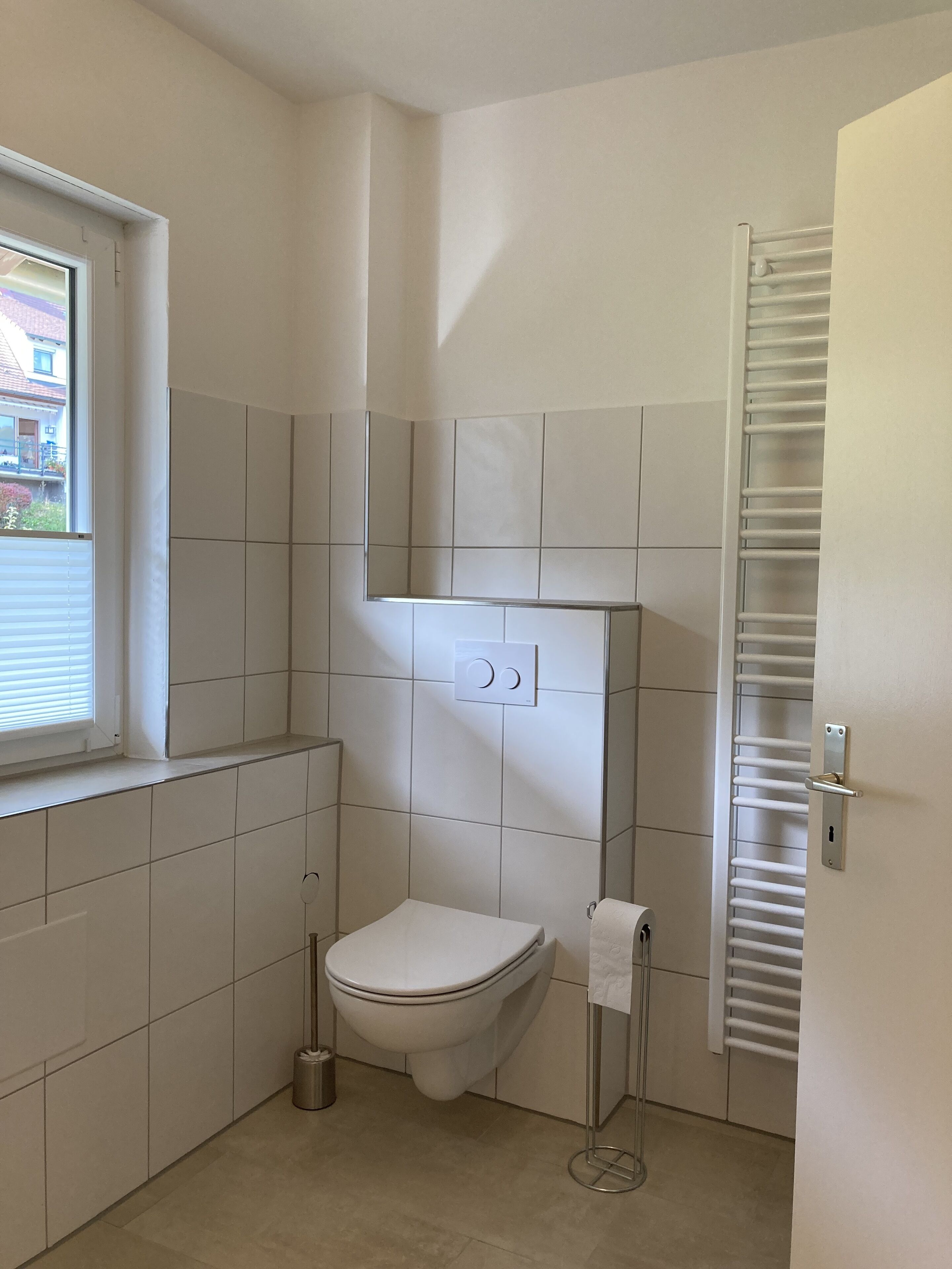 Une petite salle de bain propre, avec des carreaux blancs, des toilettes, un porte-serviettes et une fenêtre avec des stores.