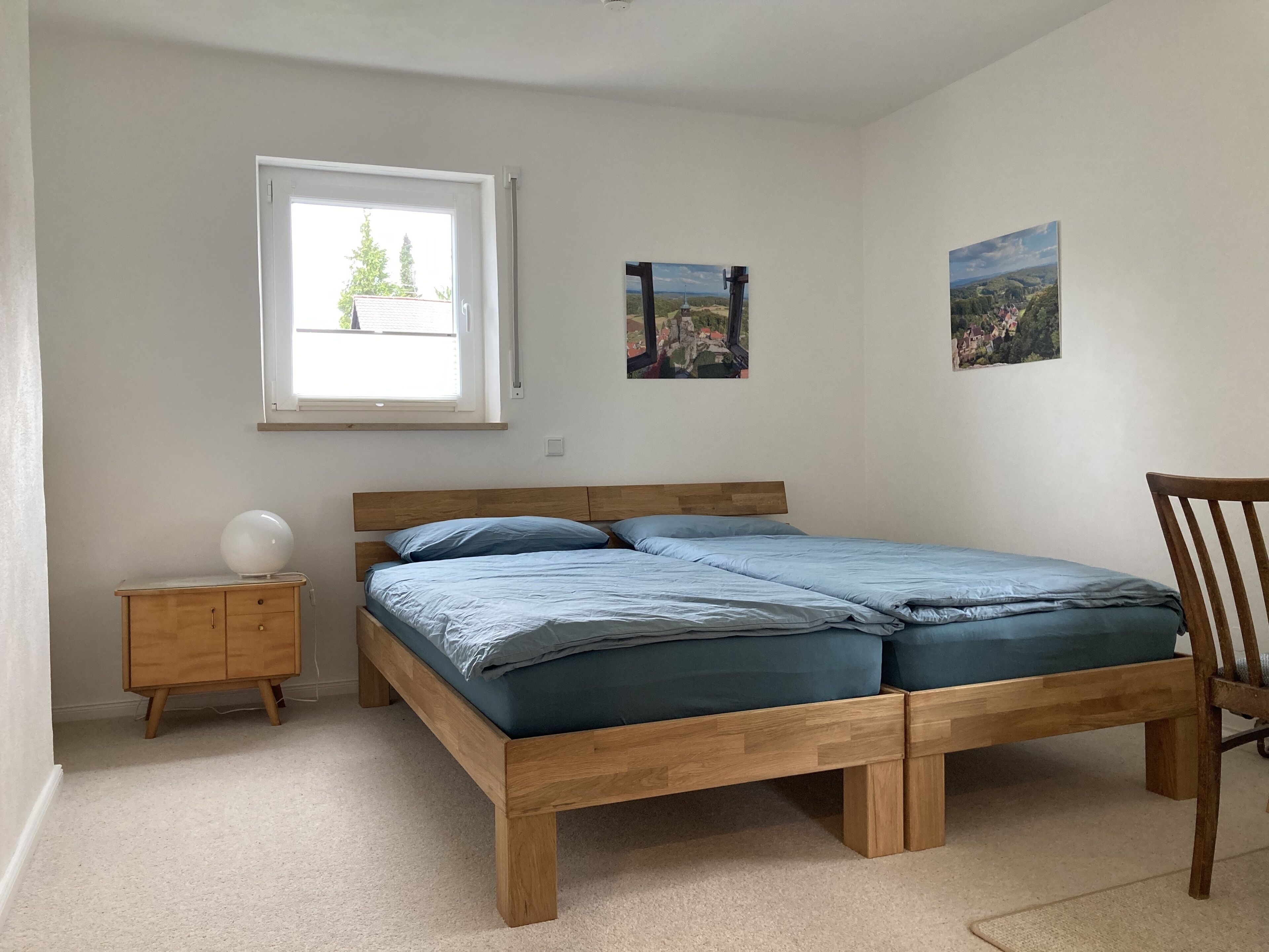 A bedroom with a wooden bed, a small bedside table, a chair, and a window with a view of trees and buildings.