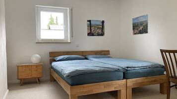 A bedroom with a wooden bed, a small bedside table, a chair, and a window with a view of trees and buildings.