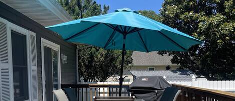 A wooden deck with a table and chairs under a blue umbrella, a white fence, and a house in the background.