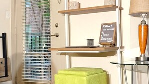 A room with a wooden floor, a green ottoman, a metal lamp, a bookshelf with decorative items, and a door with blinds. - World Cup-Ready! - Walk to MBS - Parking - ADA (Atlanta)