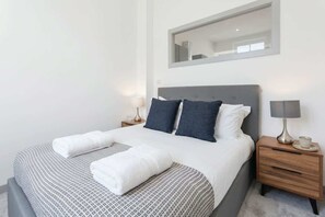 Un letto rifatto con cura, con biancheria da letto bianca e fantasia, due lampade ai lati del letto, un comodino in legno e uno specchio sopra il letto.