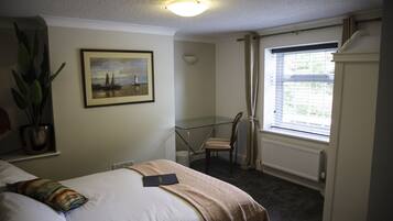 A hotel room with a bed, a desk, a chair, a radiator, a window with curtains, a framed picture on the wall, and a potted plant.