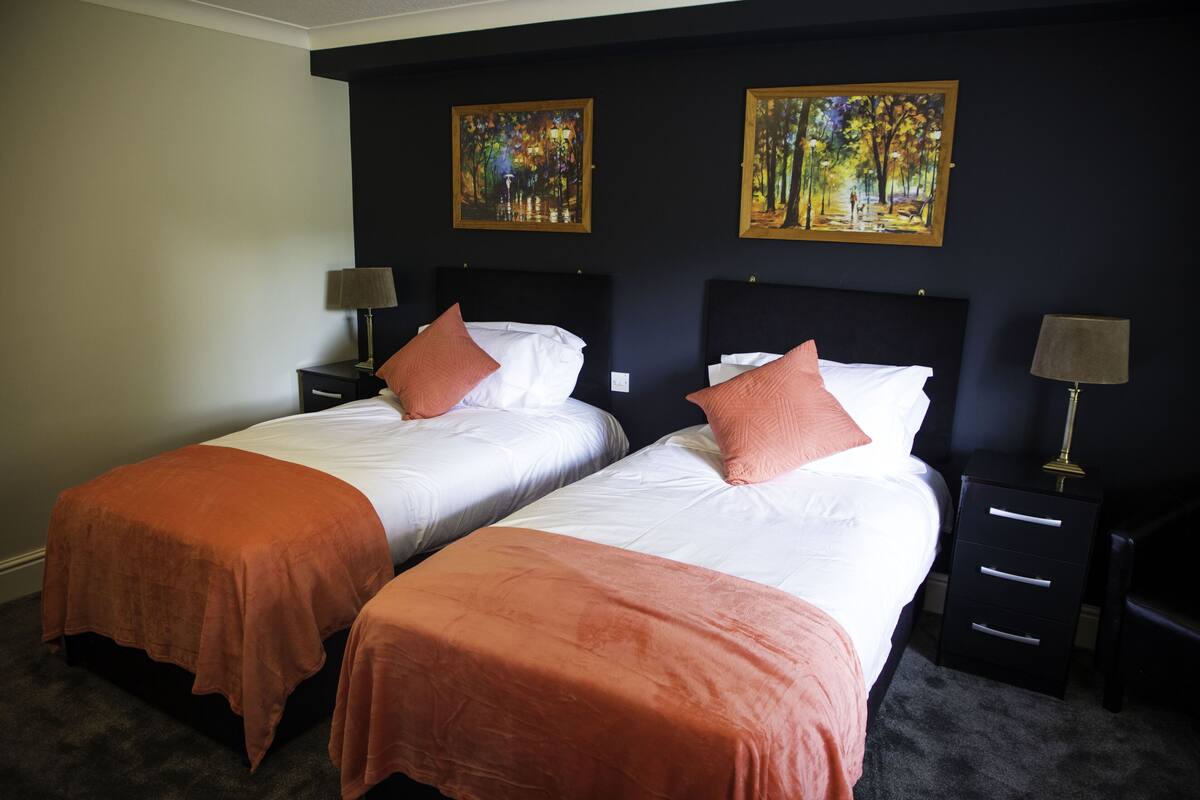 Ein Hotelzimmer mit zwei Betten, jedes mit einer weißen Matratze und einer orangefarbenen Bettdecke. Es gibt zwei Nachttische mit Lampen, und die Wände sind dunkelblau gestrichen.