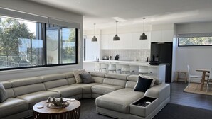 A modern living room with a L-shaped sectional sofa, a round coffee table, and a kitchen area with white cabinets and pendant lighting. - Luxury on Bayview Wifi Linen included (Inverloch)