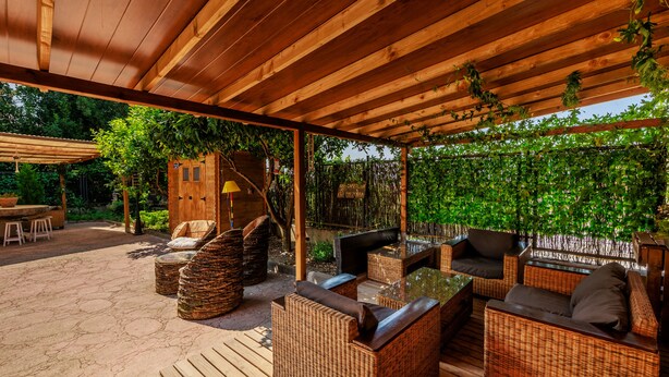 An outdoor patio with wicker furniture, a wooden pergola, and a lush green backdrop. - La Posada de la Gula (Jarandilla de la Vera)