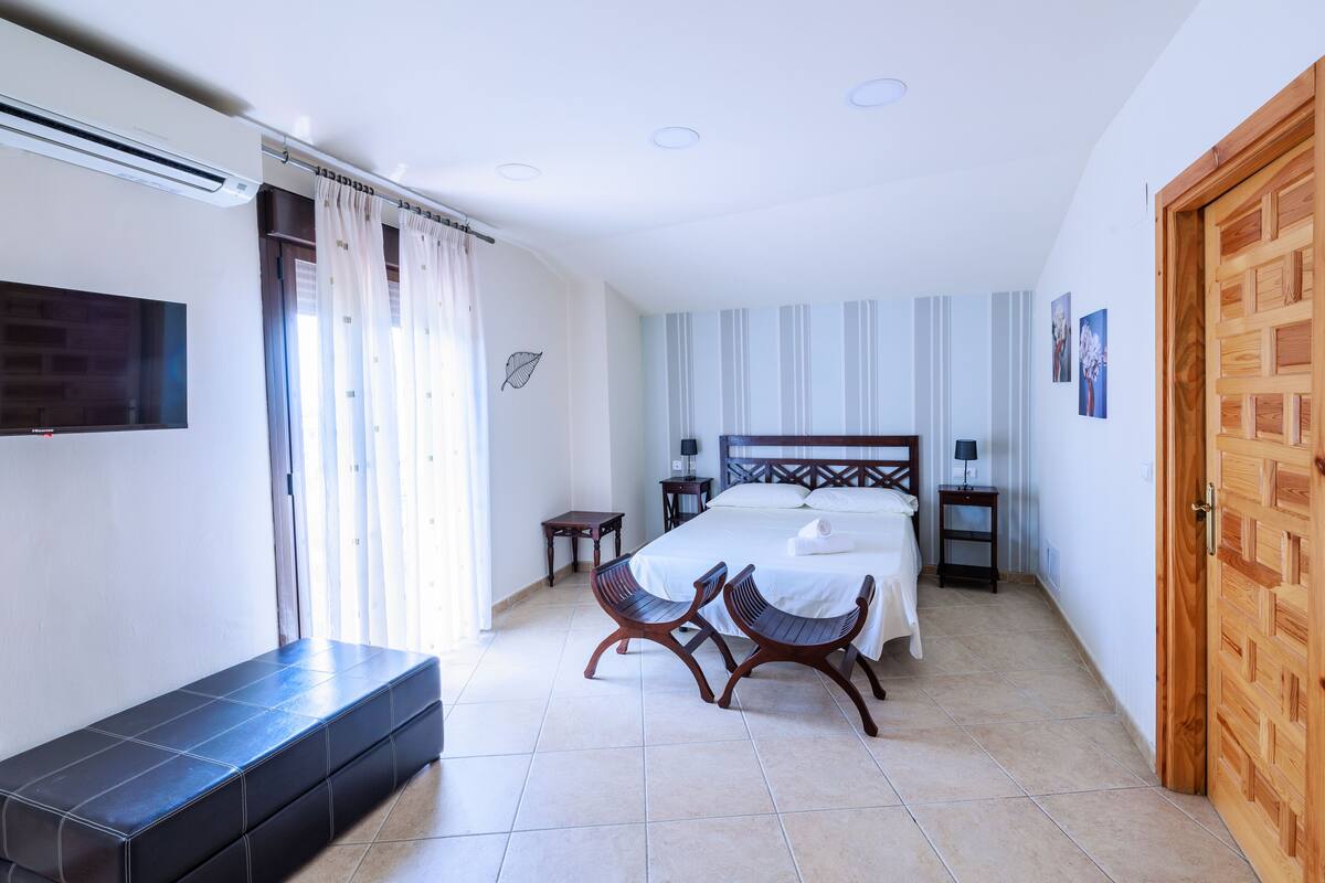 Un dormitorio con cama, dos sillas, un banco, un televisor y una puerta de madera.