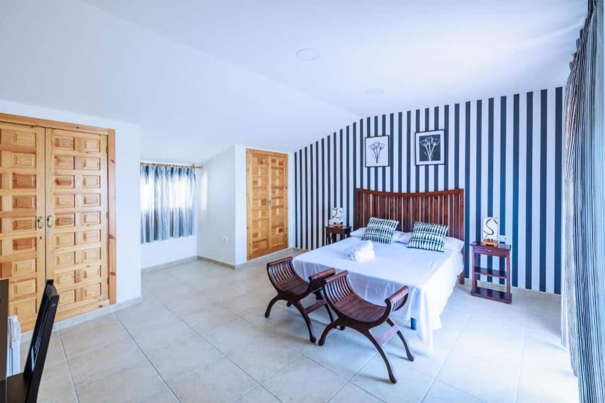 Un dormitorio con una cama grande, dos sillas de madera, una mesita de noche y un muro a rayas.