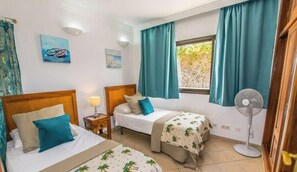 Une chambre avec deux lits, une table de chevet avec une lampe, un ventilateur et une fenêtre avec des rideaux.