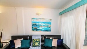 Un soggiorno con un divano in pelle nera, cuscini color acquamarina e bianchi, un quadro raffigurante un mare blu e pareti bianche.