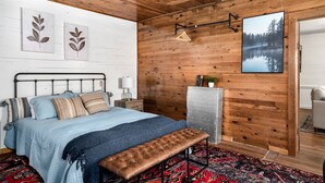 A bedroom with a wooden ceiling, a bed with a blue quilt, a bench, a wall-mounted drying rack, and a framed picture of a forest. - 2BR chalet w/ hot tub, porch & refreshing views (Mathias)