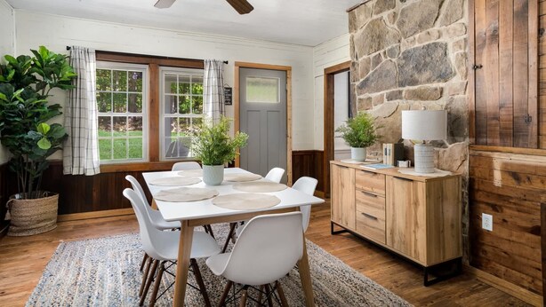 A dining area with a wooden table, white chairs, a ceiling fan, and a stone wall. - 2BR chalet w/ hot tub, porch & refreshing views (Mathias)