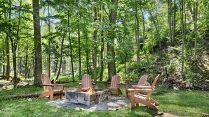 A forested area with a fire pit and wooden chairs. - 2BR chalet w/ hot tub, porch & refreshing views (Mathias)