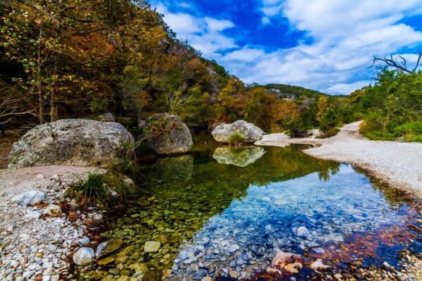A clear stream with rocks and pebbles, surrounded by trees and a clear sky.