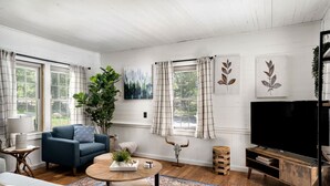 A living room with a blue sofa, a coffee table, a TV stand, and a large plant. - 2BR chalet w/ hot tub, porch & refreshing views (Mathias)