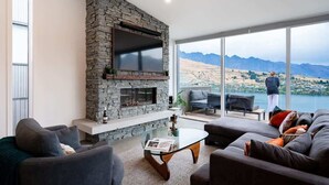 A modern living room with a stone fireplace, a glass coffee table, and a view of mountains and a lake. - Toroa Chalet| Hot Tub, Gas Fire, BBQ, Free Parking (Queenstown)