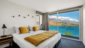 A hotel room with a large bed, a nightstand, a desk, and a view of a mountainous landscape through a sliding door. - Toroa Chalet| Hot Tub, Gas Fire, BBQ, Free Parking (Queenstown)