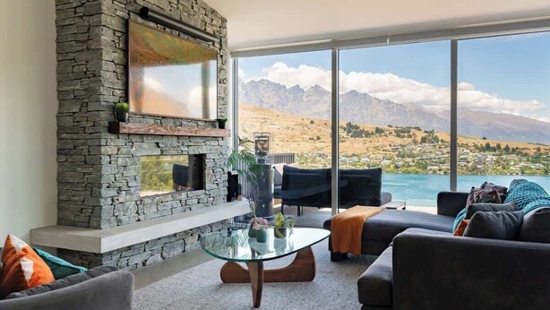 A living room with a stone fireplace, a glass coffee table, and a view of mountains and a lake. - Toroa Chalet| Hot Tub, Gas Fire, BBQ, Free Parking (Queenstown)
