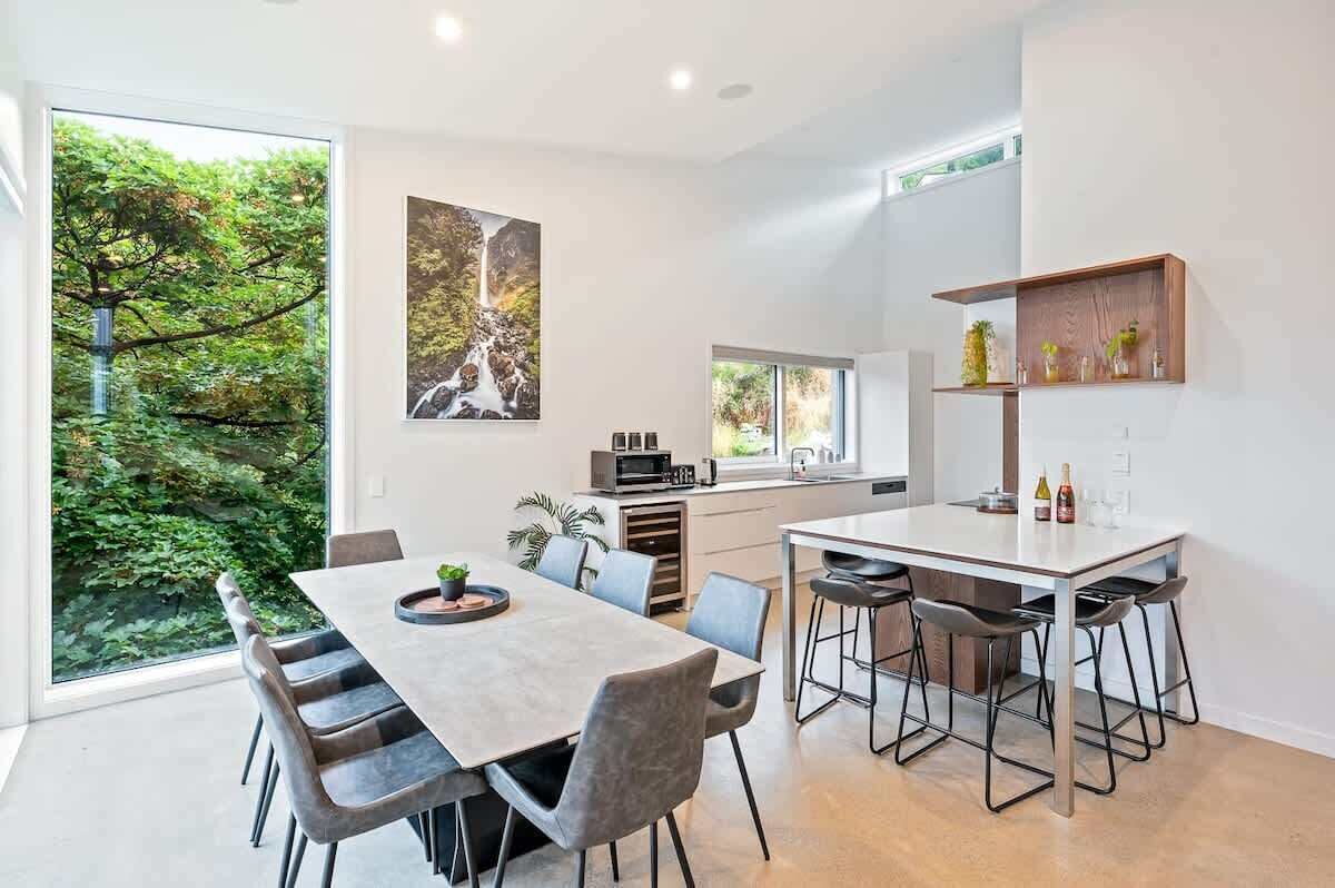 A modern kitchen with a dining area, large windows, and a view of greenery.