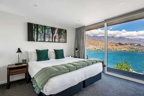 Une chambre avec un grand lit, une table de chevet, une lampe, un tableau et une vue sur un paysage montagneux, visible à travers une porte vitrée.