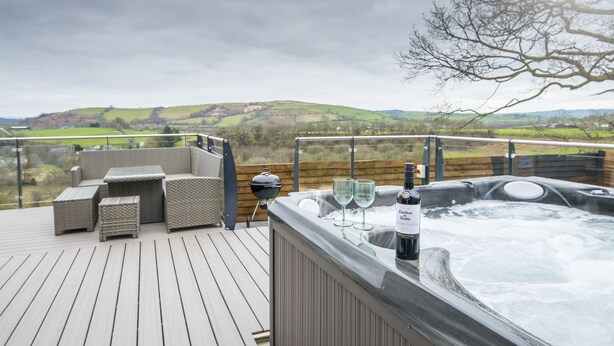 A hot tub on a deck with wicker furniture and a view of rolling hills. - Luxury Accessible Holiday Home with Hot Tub & Views (Llansawel)