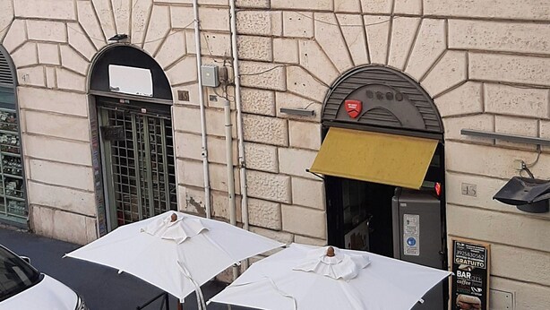 An outdoor cafe with white umbrellas, a stone building, and a parked car. - Augustus Room (Roma)