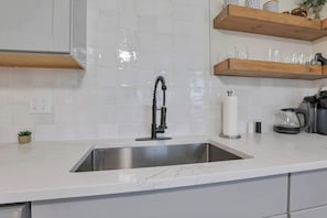Una cocina con un lavamanos de acero inoxidable, encimera de mármol blanco, estanterías de madera y una pequeña planta en maceta.