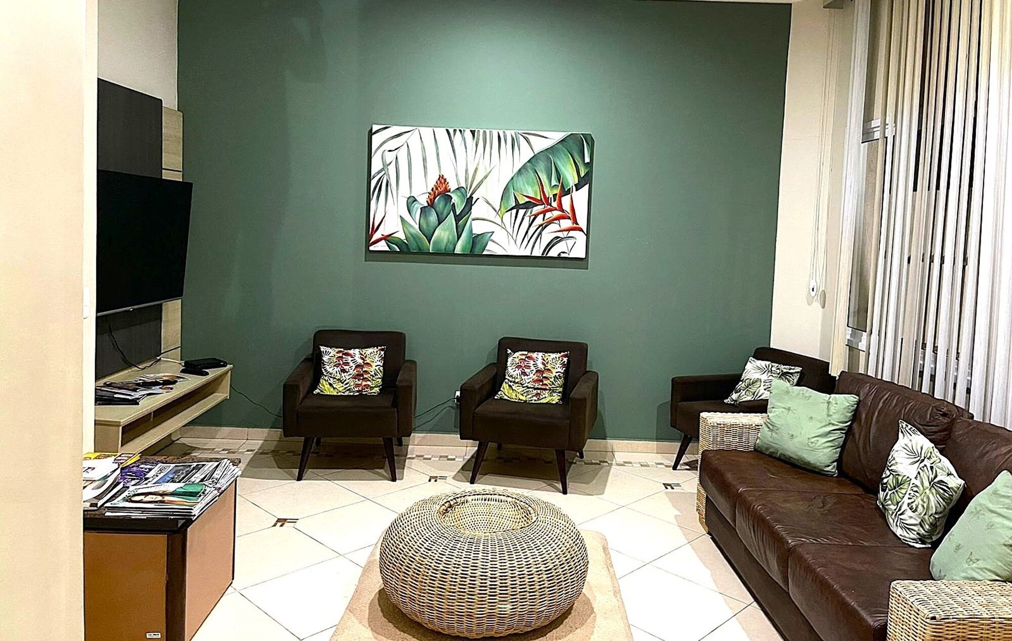 A living room with a green wall, two armchairs with patterned cushions, a brown sofa with cushions, a round woven ottoman, a flat-screen TV, and a small table with magazines.