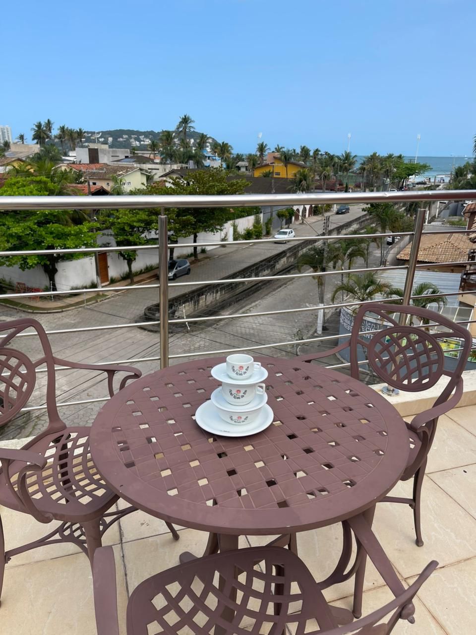 Mesa redonda con un juego de té en un balcón con vistas a una zona costera con palmeras y edificios.