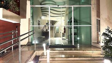 Entrada de hotel con fachada de vidrio, una escalera y barandilla metálica.