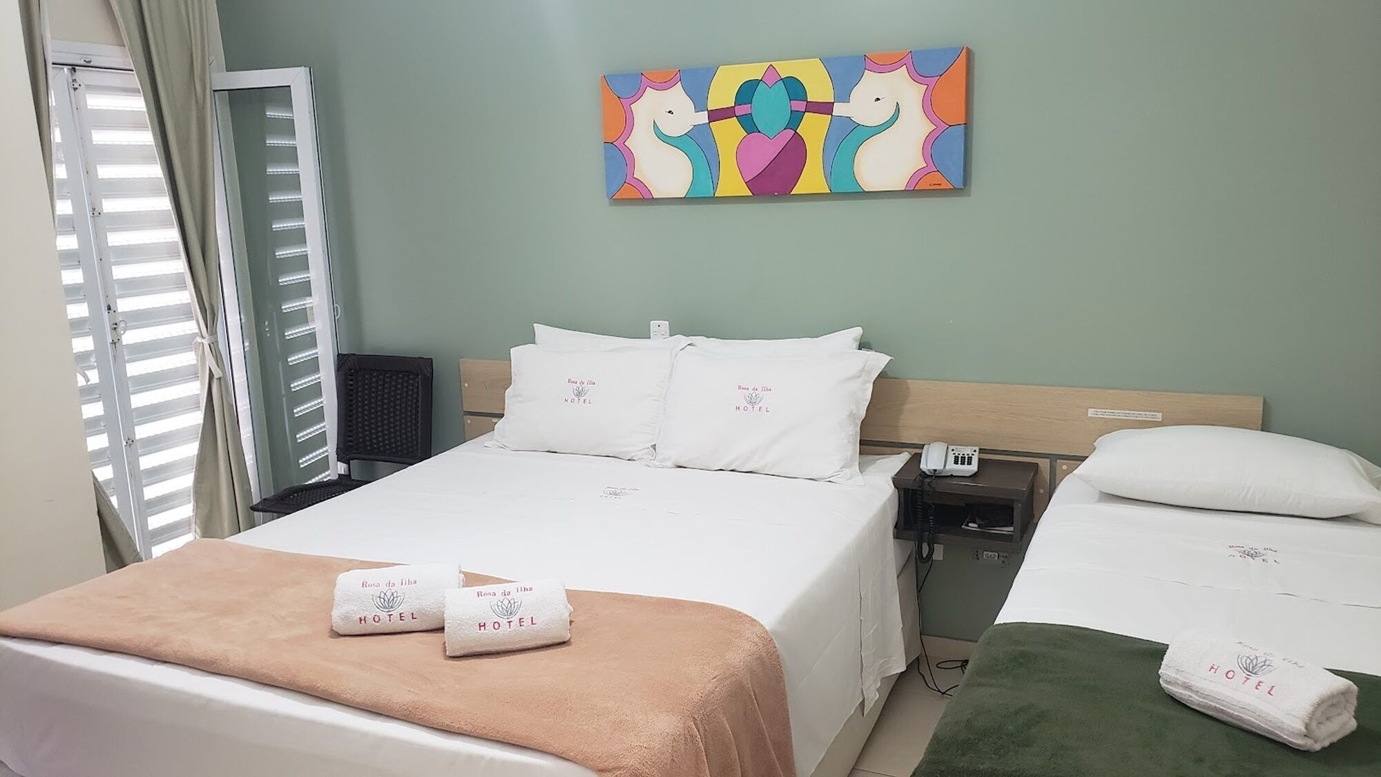 Une chambre d’hôtel avec un lit, deux tables de chevet, une commode, un tableau et une fenêtre avec des stores.