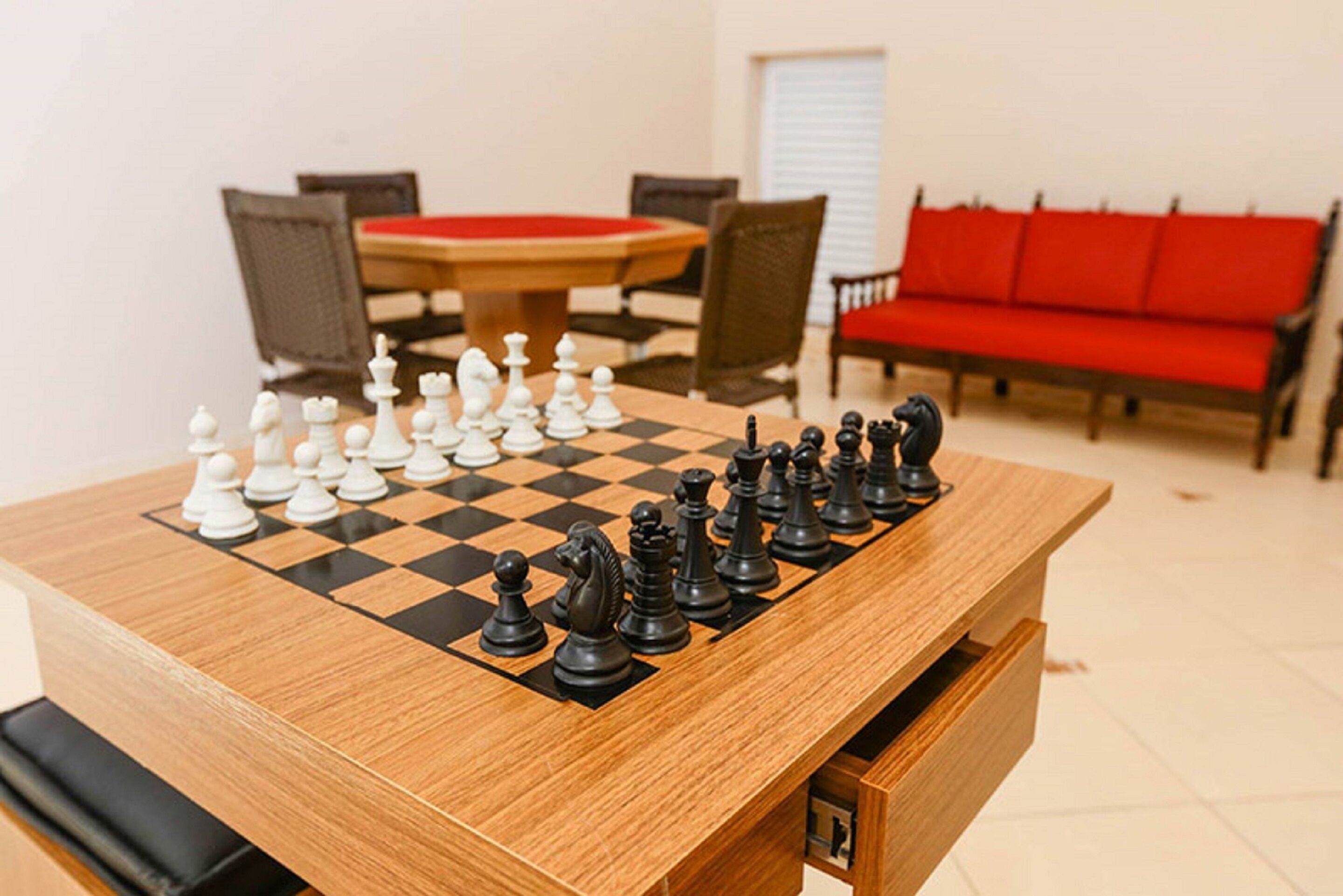 A chessboard set up on a wooden table in a room with a red sofa and a dining table in the background.