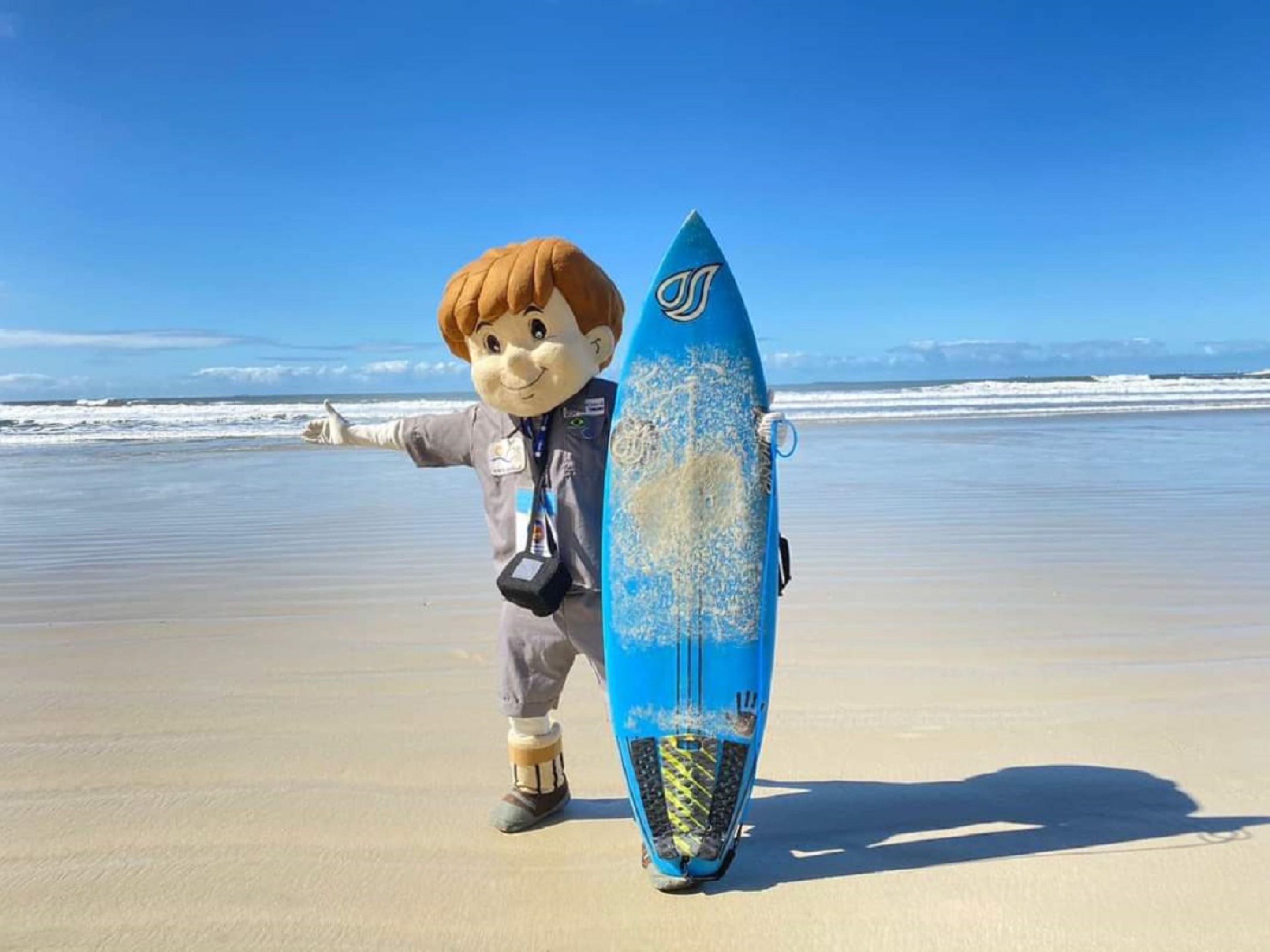Une mascotte tenant une planche de surf bleue sur une plage de sable.