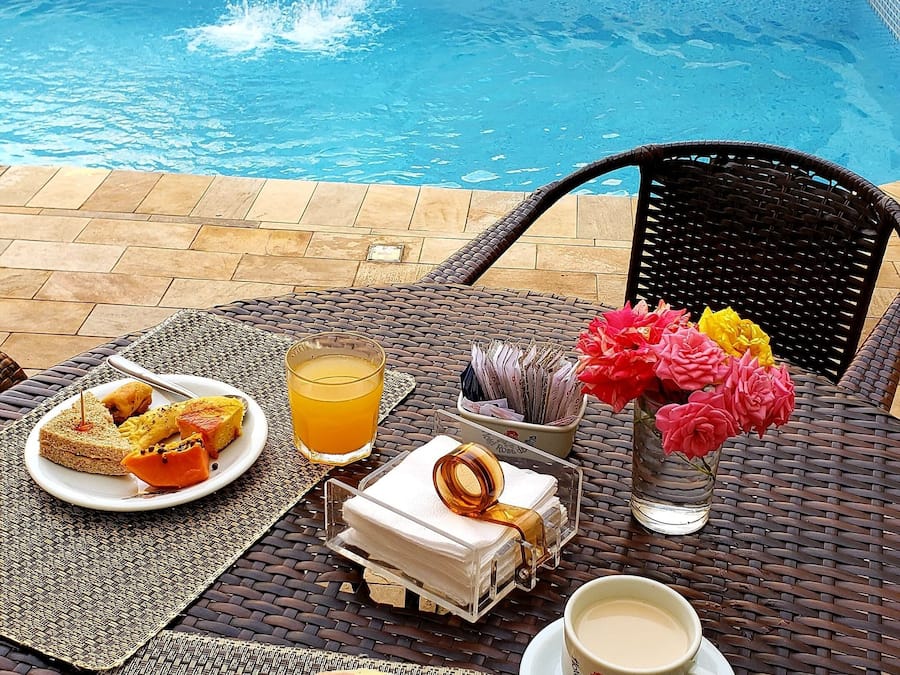 Una mesa de comedor con alimentos para el desayuno, un vaso de zumo de naranja y una taza de café, con vista a la piscina.