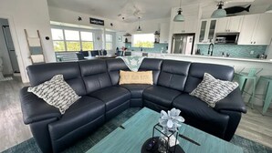 Sala de estar moderna com um sofá modular de couro preto, uma mesa de centro verde-água e uma cozinha ao fundo.