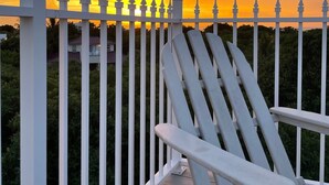 Uma cadeira de deck branca em um deck de madeira com corrimão branco, com vista para um pôr do sol com tons laranja e amarelos no céu.