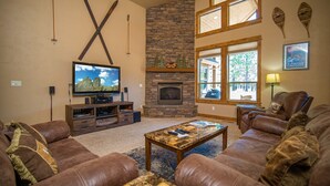 A living room with a stone fireplace, leather sofas, a flat-screen TV, and large windows. - Sunriver Forest Escape with Access to Forest House (Sunriver)