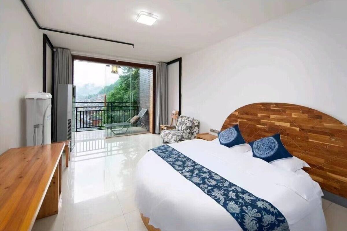 Een slaapkamer met een groot bed, een houten hoofdbord, een houten bankje, een klein tafeltje en een balkon met uitzicht op groen.