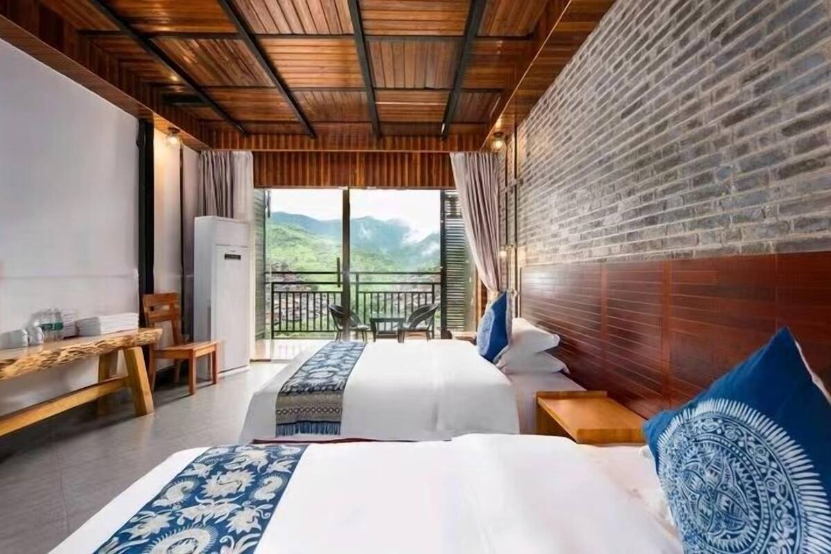 Een hotelkamer met twee bedden, een houten plafond, een bakstenen muur en een balkon met uitzicht op bergen.