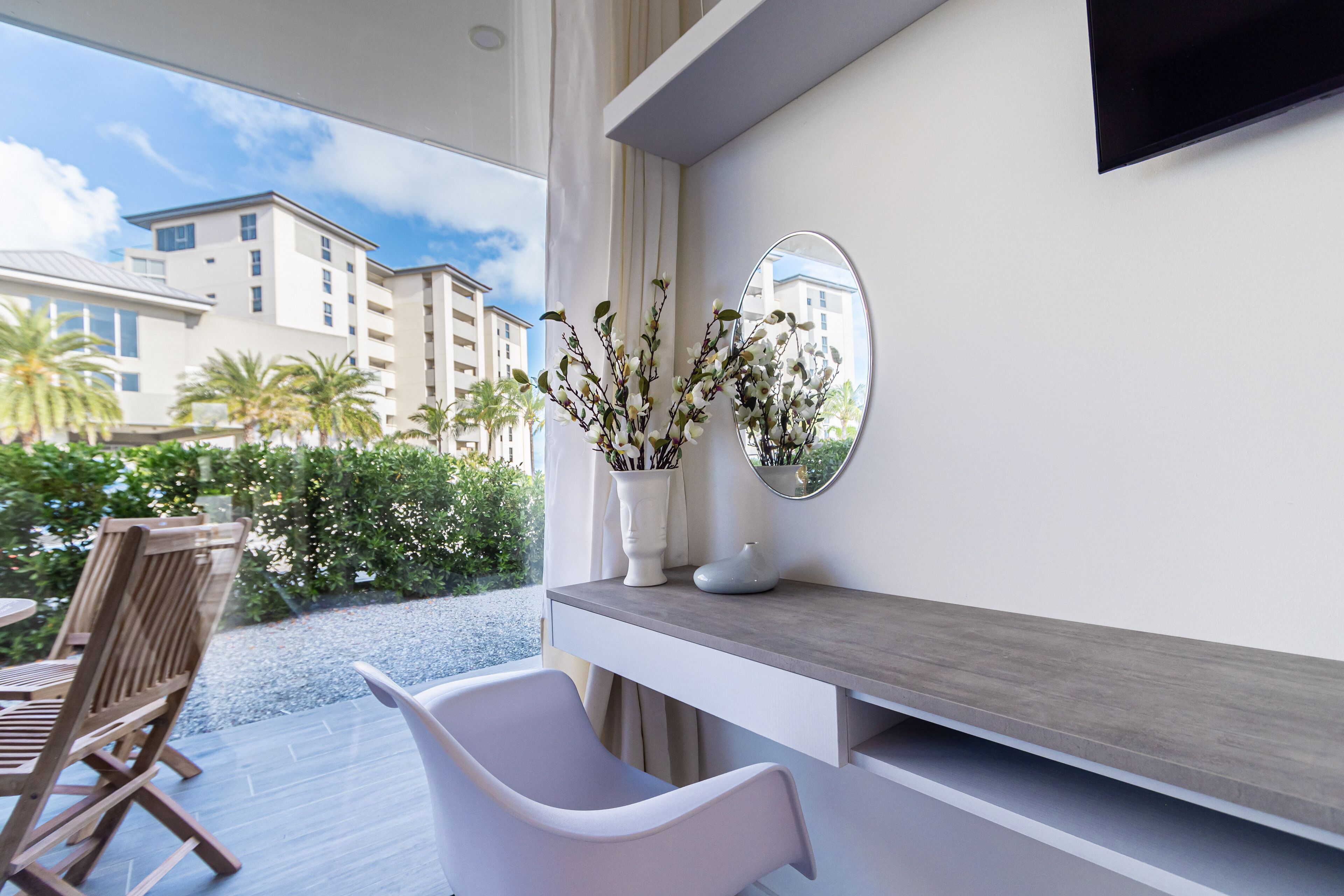 A modern room with a large window, a wooden chair, a white chair, a grey console table, and a mirror.