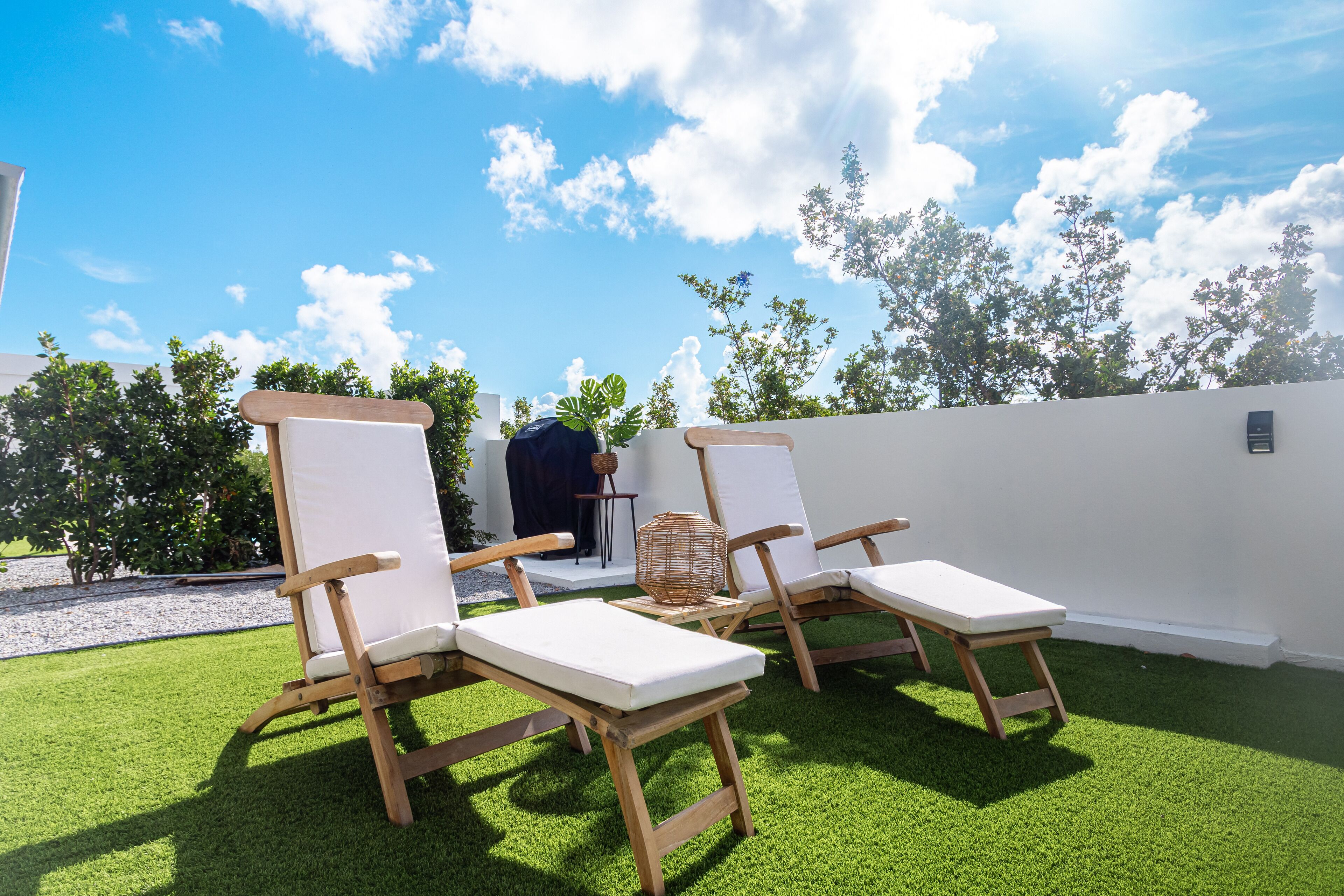 Un patio con dos sillones de madera y una pequeña mesa, rodeado por un muro blanco y césped artificial verde.
