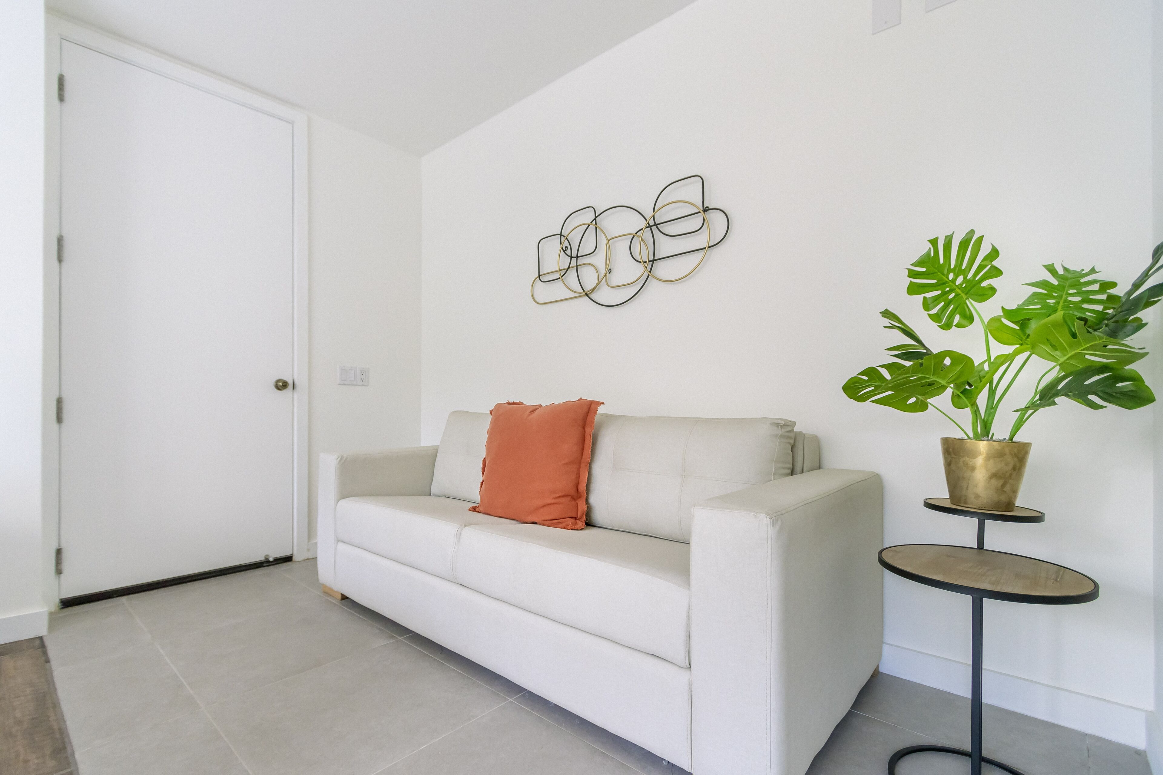 A white sofa with a single orange pillow, a round side table with a potted plant, and a wall art piece.