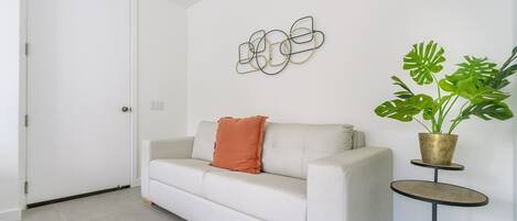 A white sofa with a single orange pillow, a round side table with a potted plant, and a wall art piece.