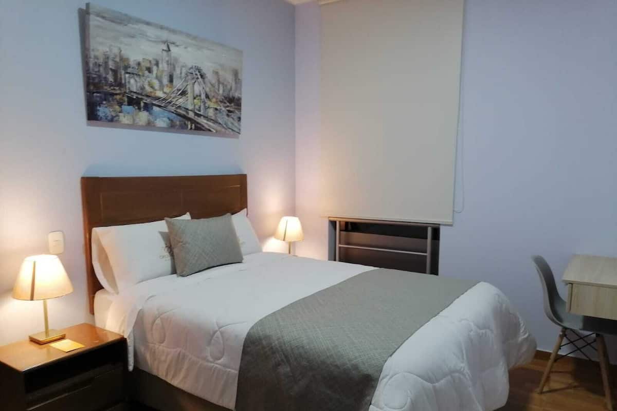 A neatly made bed with a wooden headboard, two bedside tables with lamps, a painting on the wall, and a window with a blind.