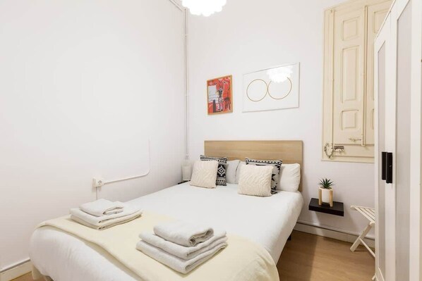 Un lit bien fait, avec du linge de lit blanc et une serviette pliée, une tête de lit en bois, une petite étagère avec une plante et une œuvre d’art accrochée au mur.