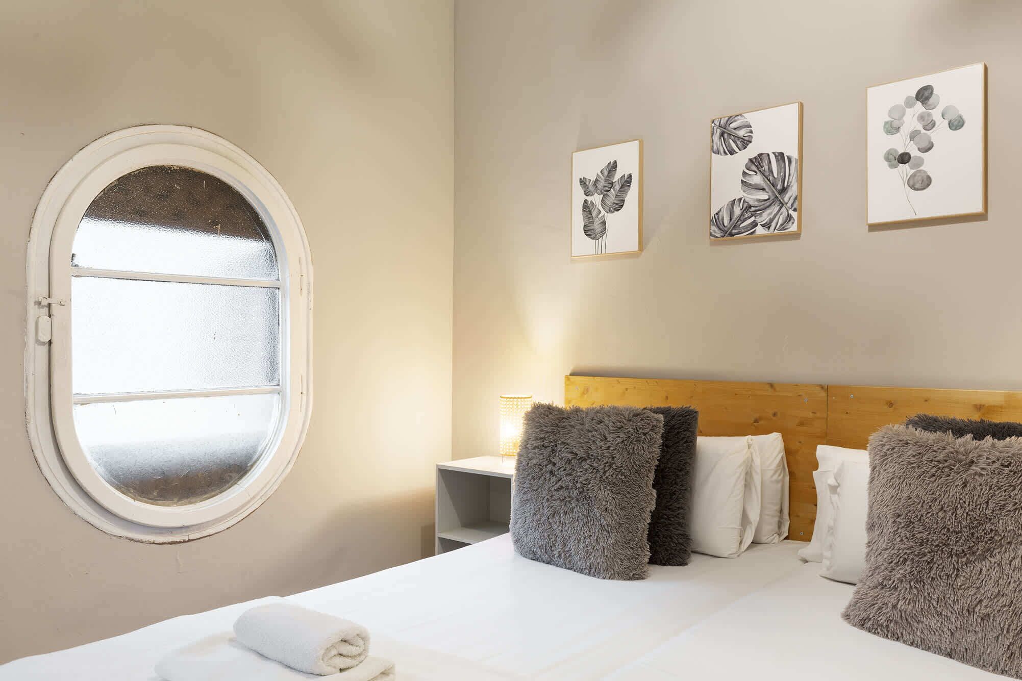 Ein Hotelzimmer mit einem Rundfenster, einem hölzernen Kopfteil und drei gerahmten Pflanzenbildern an der Wand.