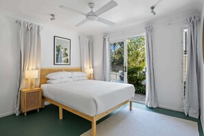 Une chambre avec un lit, deux tables de chevet avec des lampes, un ventilateur de plafond, un tableau encadré au mur et une vue sur la verdure à l’extérieur.