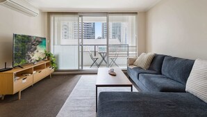 A modern living room with a grey sofa, a wooden TV stand, and a flat-screen TV. There is a balcony with a table and chairs, and a city view through large windows. - Gorgeous 3-Bed Apartment Near Flagstaff Gardens (West Melbourne)