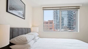 A neatly made bed with white linens and pillows, a gray upholstered headboard, a bedside lamp, and a window with a view of a modern building and a brick building. - Gorgeous 3-Bed Apartment Near Flagstaff Gardens (West Melbourne)